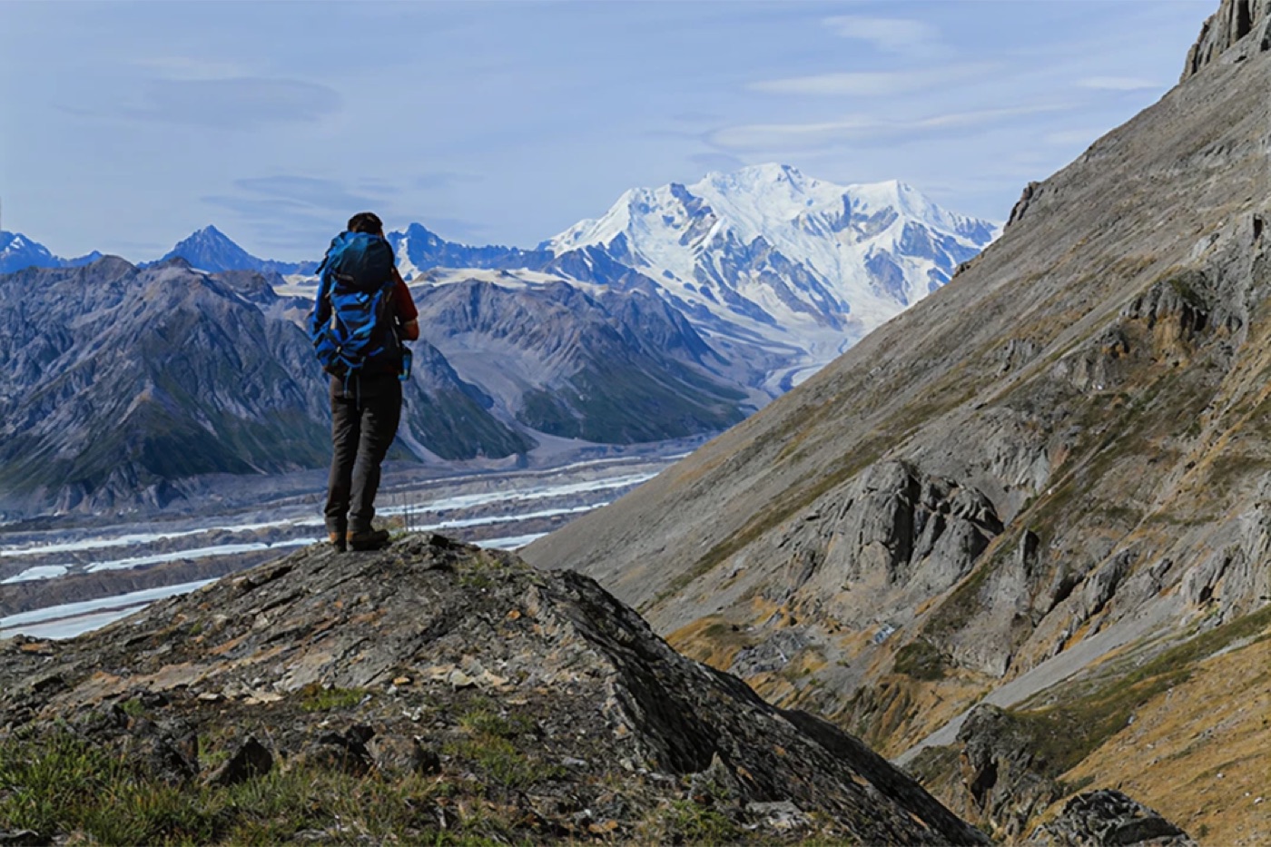 Hiker with a backpack on a ridge facing snow-capped mountains and a glacier valley
